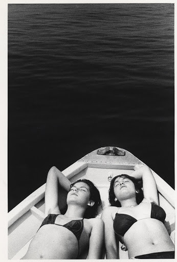 Two women in bikinis lie on a boat under the sun, black and white photograph, calm water, carefree summer moment, leisure and relaxation.