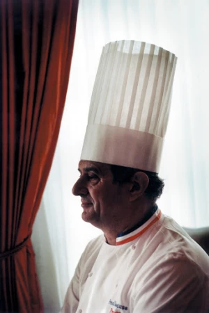 Profile portrait of an adult male chef wearing a toque, seated near a window with red curtains, side lit indoors.