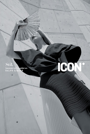Fashion editorial shot in black and white. A model poses with a fan in front of a geometric concrete wall with stylized typography.