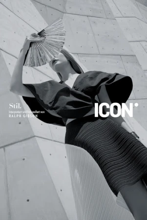 Fashion editorial shot in black and white. A model poses with a fan in front of a geometric concrete wall with stylized typography.