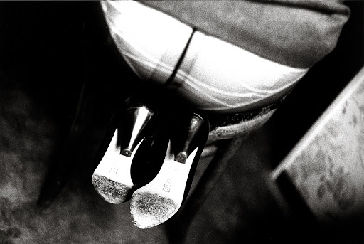 High angle black and white shot of women's high heels with their soles facing up, next to a seated person in patterned fabric.