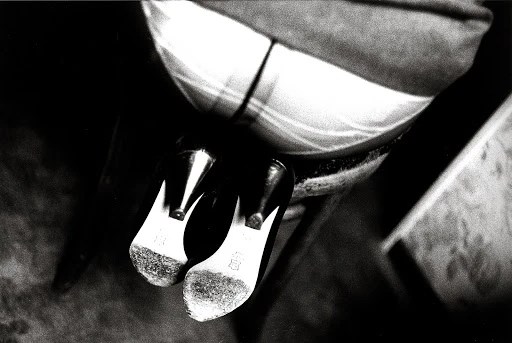 High angle black and white shot of women's high heels with their soles facing up, next to a seated person in patterned fabric.