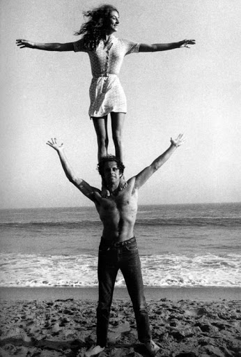 A man stands on a sandy beach balancing a woman on his shoulders as both extend their arms outward.