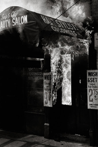 Black and white photo of a hair salon with smoke, featuring signs for haircuts and wigs, evoking a vintage or documentary style.