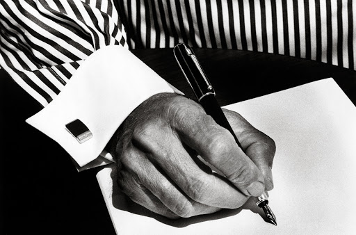 Close-up of an adult hand writing on paper with a fountain pen, wearing a striped shirt and cuff links in black and white.
