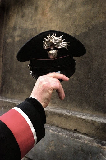 A hand holds a black Carabinieri hat with a silver emblem against a textured background, featuring a red stripe on a dark sleeve.
