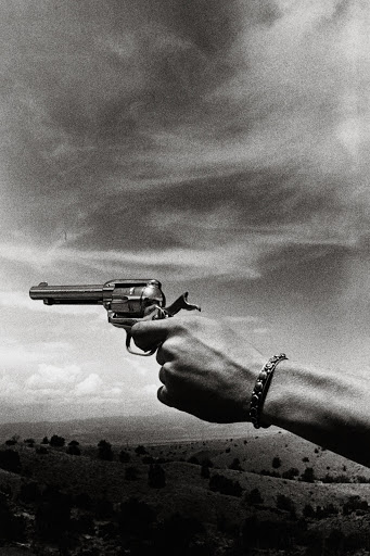 Monochrome, high-angle shot of an adult's hand holding a gun against cloudy sky and distant landscape; jewelry present on wrist.