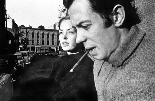 Black and white photograph of a couple outside; a man with a cigarette and a woman are in focus with an urban street behind them.
