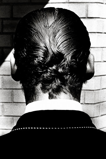 From the back, a person with styled dark hair stands near a brick wall in a black and white image with strong directional lighting.