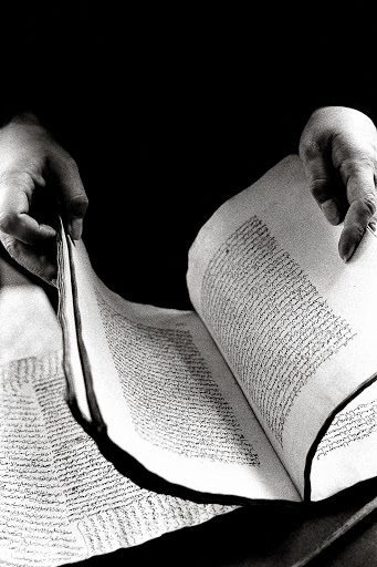 Hands holding open an antique book with Arabic or Farsi script in black and white, creating a moody and mysterious atmosphere.
