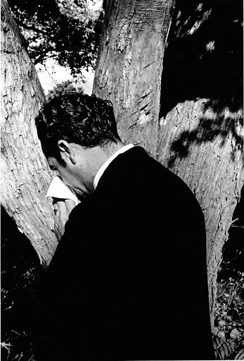 Monochrome portrait of a man in a suit blowing his nose with a tissue, standing close to a tree in an outdoor setting with natural lighting.