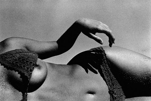Monochromatic, close-up image featuring a woman's torso in repose, accentuated by hands and textured bikini top against a plain backdrop.