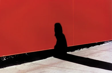 A long dark shadow of a person cast against a vibrant red wall next to a black baseboard and light concrete ground.