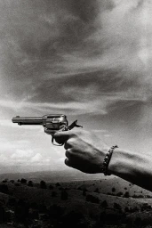 Close-up monochrome image of a hand holding a revolver against a dramatic cloudy sky and a sparse landscape by Ralph Gibson.