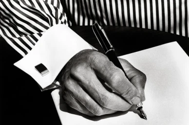 Close-up of a person's hand writing with a fountain pen on paper, wearing a striped shirt with French cuffs and cufflinks.