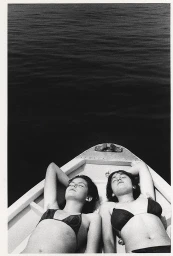 Two women lying in bikinis on the bow of a boat with dark water in the background by Ralph Gibson.