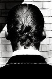 Close-up back of head of person with slicked dark hair and white shirt collar, photographed in high-contrast monochrome.