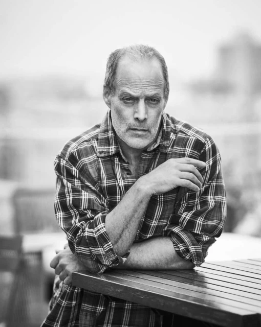 Black and white portrait of a mature adult man in a flannel shirt, leaning on a wood table outdoors with a neutral facial expression.