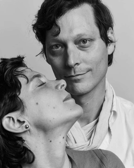 Monochrome portrait of a man and a woman, close together, with the man in a white shirt and the woman in a dark top in a studio setting.