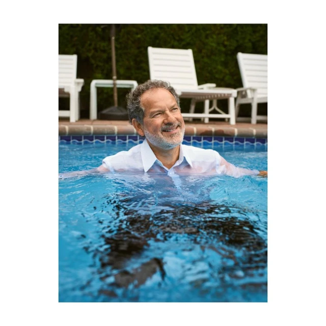 Man with gray hair smiles while standing chest-deep in a bright blue outdoor swimming pool on a sunny day, wearing a white dress shirt.
