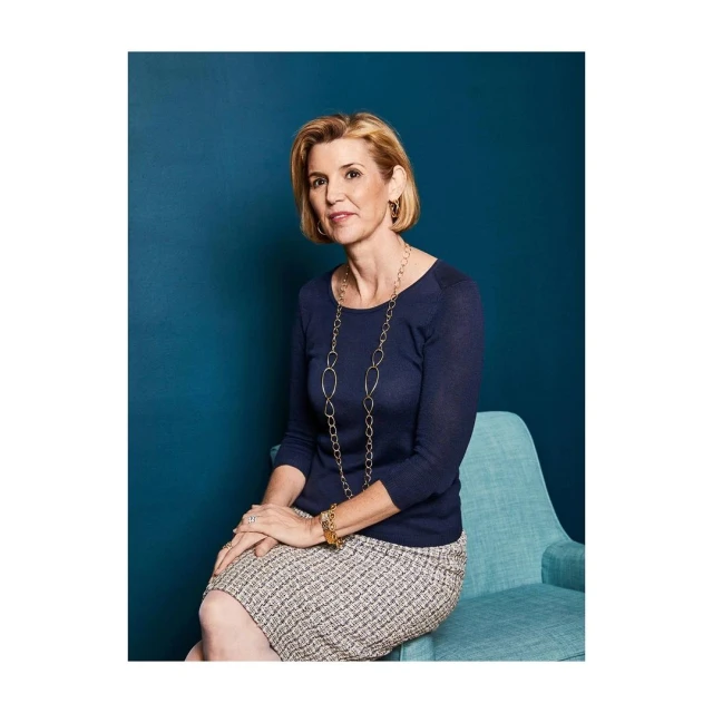 Blond woman poses indoors in blue top and tweed skirt, seated on teal chair against a dark teal wall, wearing gold jewelry.