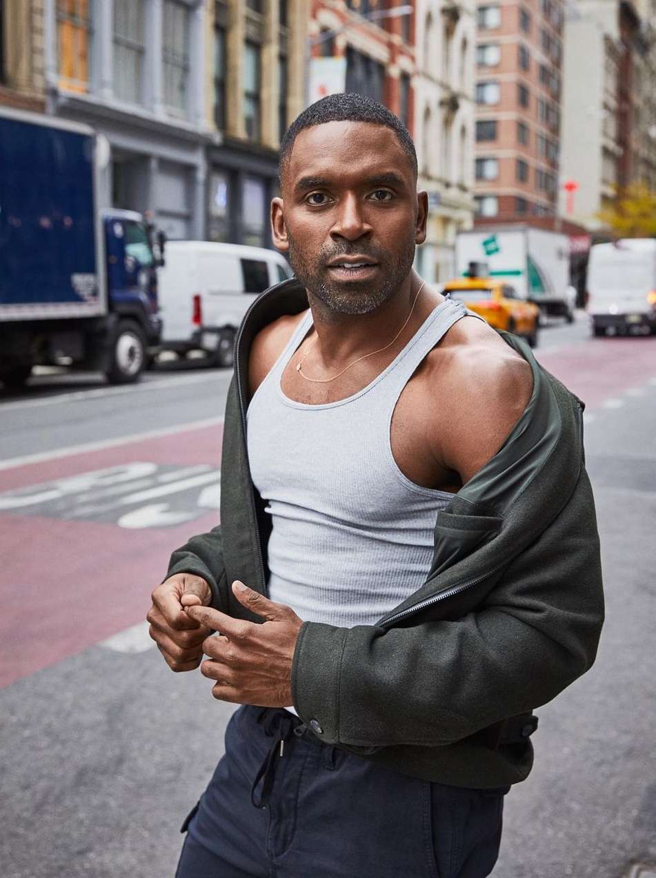 Portrait of a dark-skinned man in a white tank top and black jacket standing on a city street with buildings and vehicles in background.