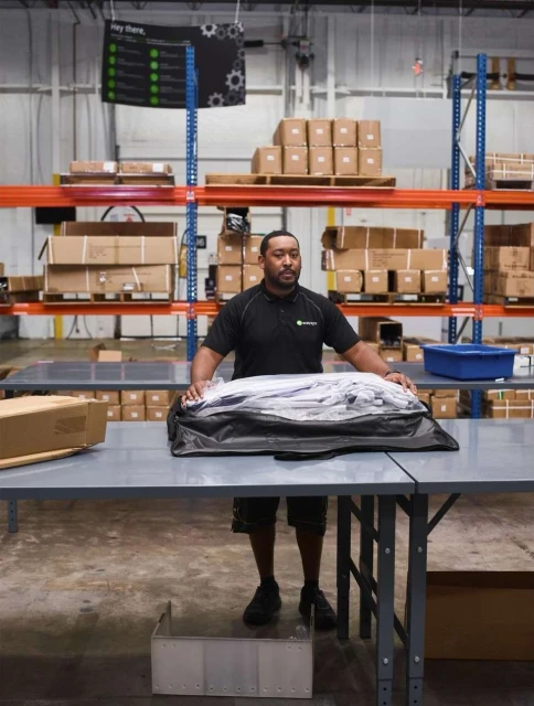 Adult man stands at a gray table in a warehouse surrounded by boxes on shelving. He wears a dark shirt and black shoes.
