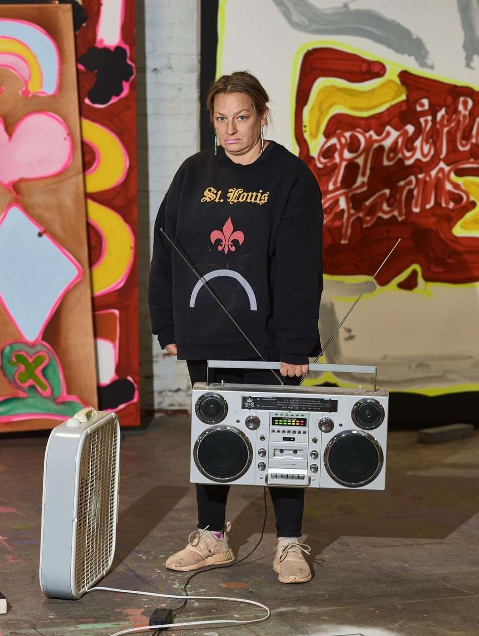Adult woman in black sweatshirt with boombox stands in front of paintings, fan plugged into an outlet on the floor, indoor environment.
