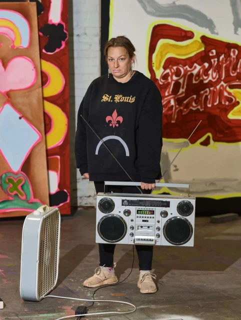 Adult woman in black sweatshirt with boombox stands in front of paintings, fan plugged into an outlet on the floor, indoor environment.