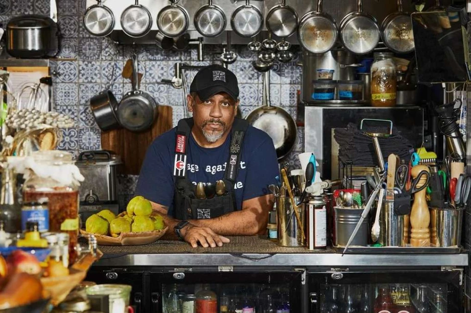 Portrait of a middle-aged African American man in a kitchen setting with hanging pots and various kitchenware, wearing a hat and apron.