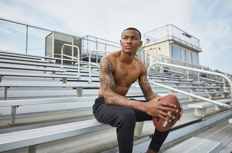 Shirtless man with tattoos sits on stadium bleachers, holding a football, and gazes thoughtfully into the distance on a cloudy day.