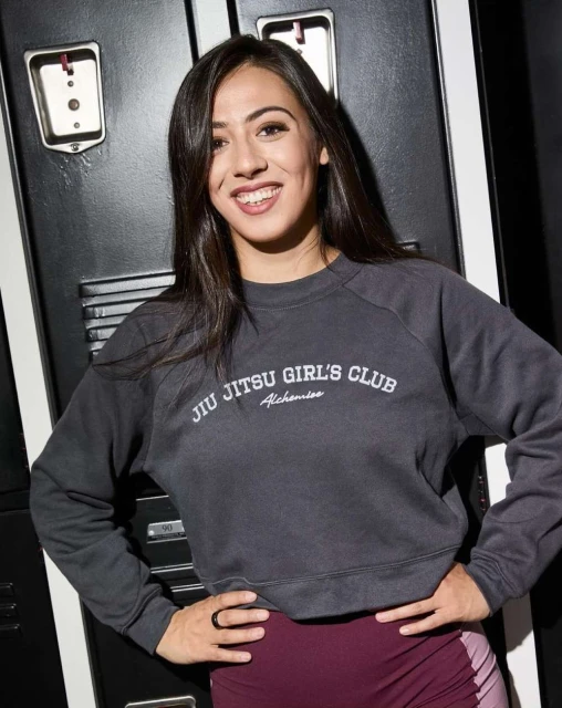 Smiling woman wearing a 'Jiu Jitsu Girl's Club' sweatshirt posing in front of gray lockers, showcasing athletic wear and casual fashion.