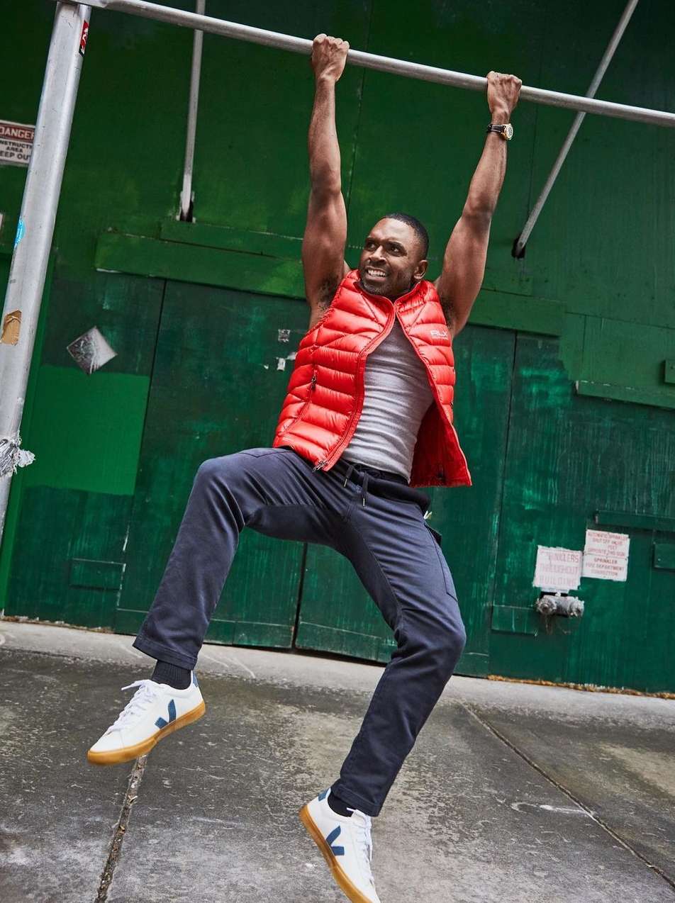 Adult man performs pull-up on outdoor bar. He wears an orange vest, grey shirt, blue pants and Veja sneakers in an urban setting.