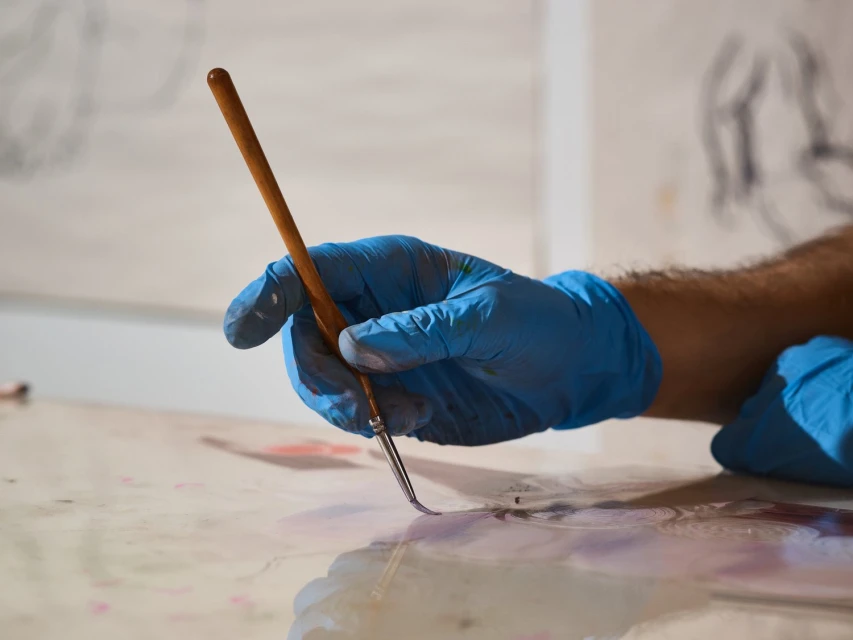 Close-up of artist's gloved hand holding fine brush over canvas with paint, set against blurred background of artwork in an indoor art studio.