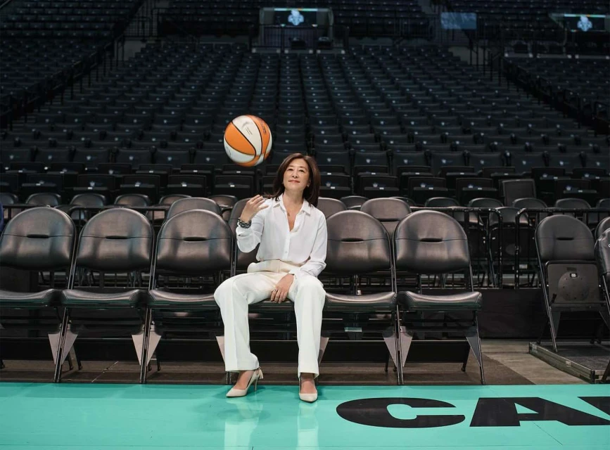 Asian woman in white suit sits on black chairs in basketball arena tossing basketball in the air with empty stadium seating background.