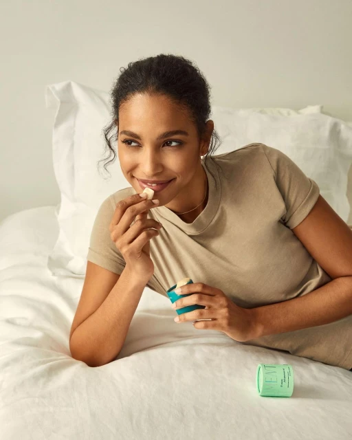 A woman in a beige shirt lies in bed holding a lozenge and a green container, relaxing in a brightly lit, minimalist bedroom setting.