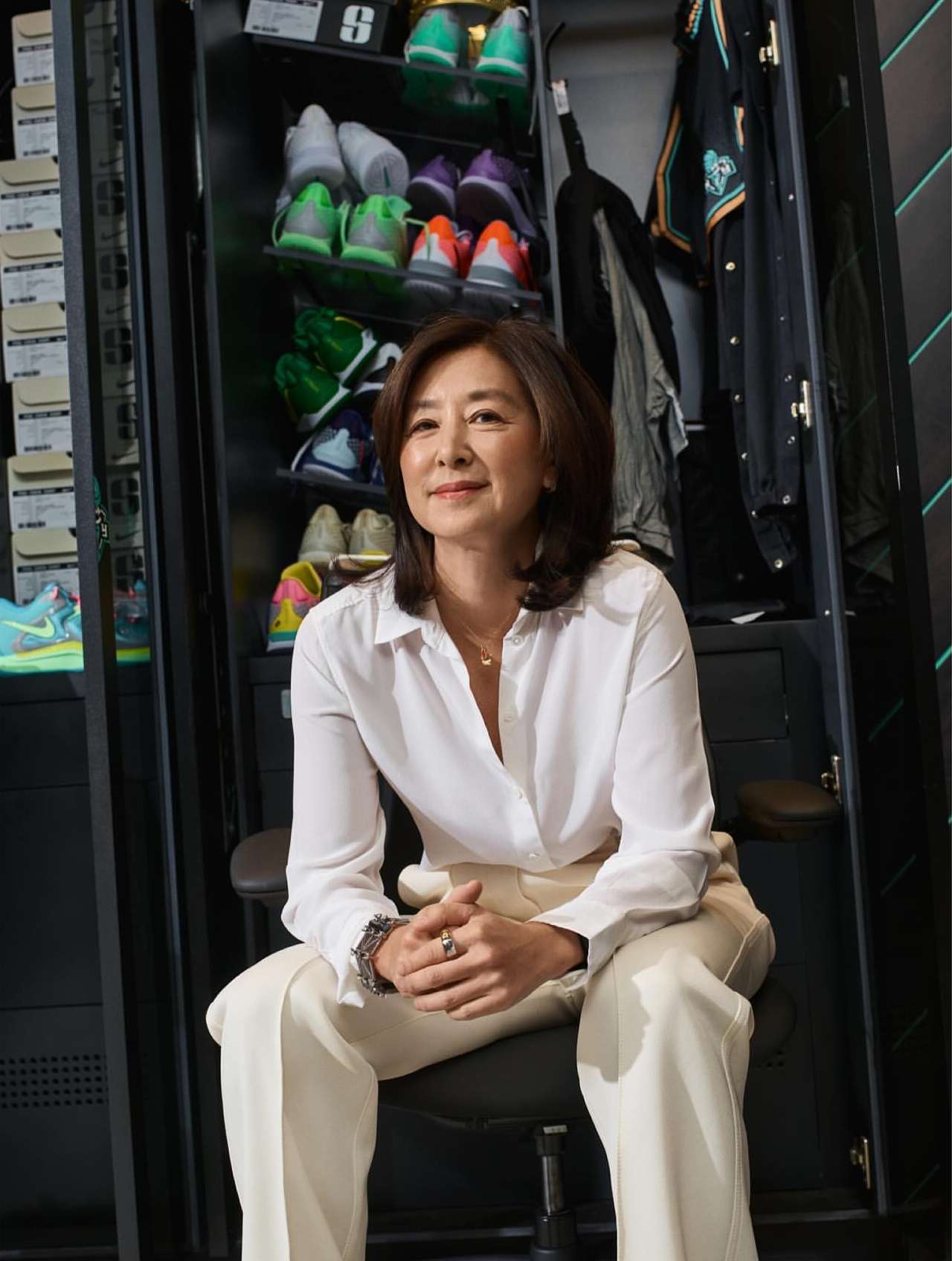 An adult woman sitting on a chair in a room with a shoe locker, wearing a white blouse and light-colored pants.