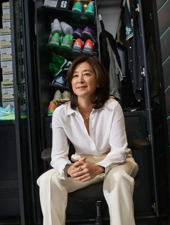 An adult woman sitting on a chair in a room with a shoe locker, wearing a white blouse and light-colored pants.