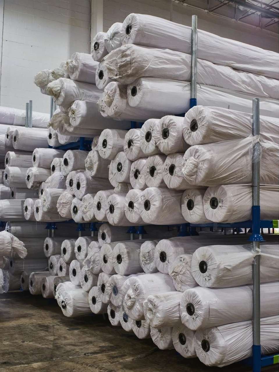 Stacks of wrapped textile rolls on a blue metal industrial shelving rack in a warehouse setting, showcasing storage and distribution.