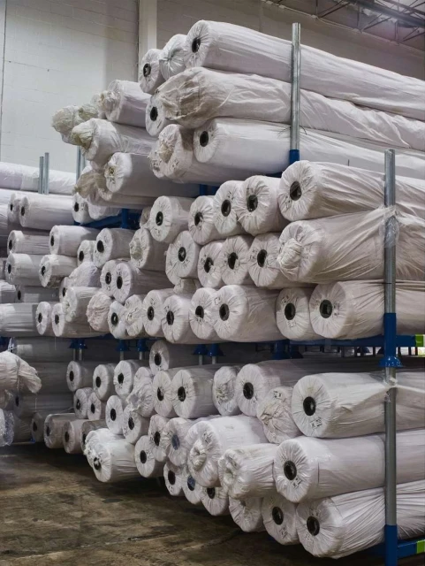 Stacks of wrapped textile rolls on a blue metal industrial shelving rack in a warehouse setting, showcasing storage and distribution.