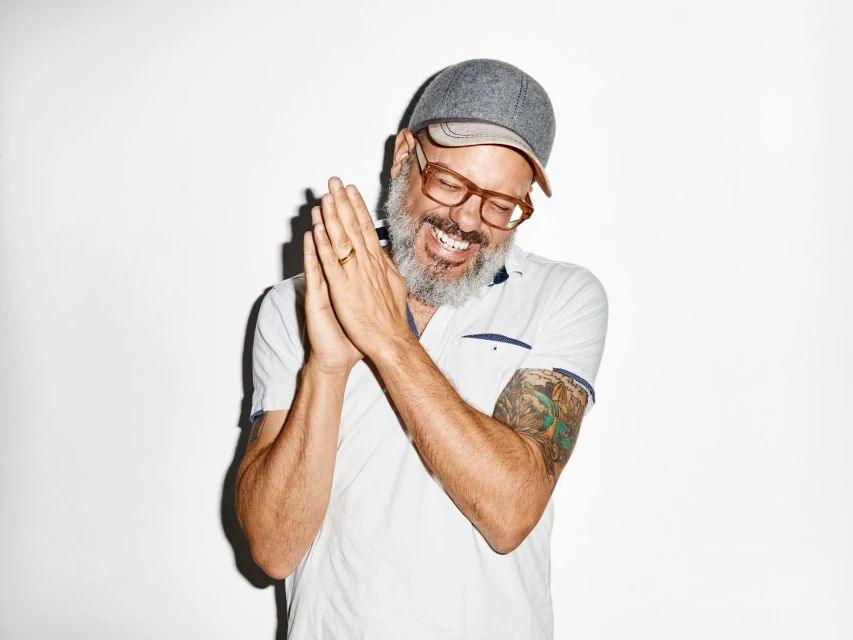 Smiling man with a beard wearing glasses and a cap, hands together, in a studio shot with a white background and a tattoo on arm.