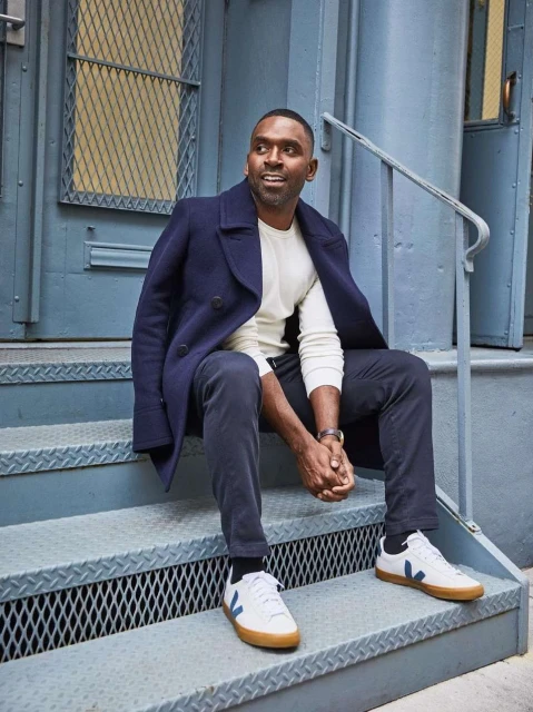 A dark-skinned man wearing a wool coat and sneakers sits on a metal staircase with a light-blue door as a backdrop in an outdoor setting.