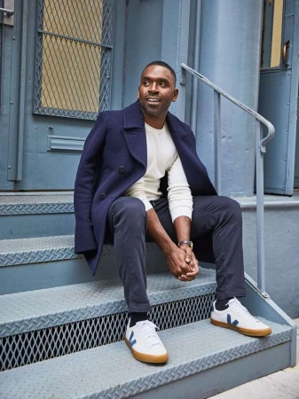 A dark-skinned man wearing a wool coat and sneakers sits on a metal staircase with a light-blue door as a backdrop in an outdoor setting.