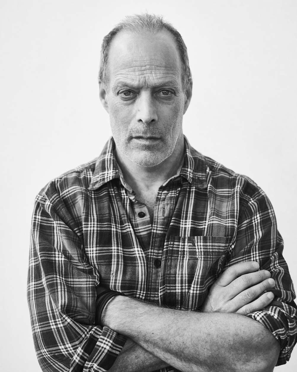Black and white portrait of a middle-aged man in a plaid shirt with crossed arms against a white background in an indoor setting.