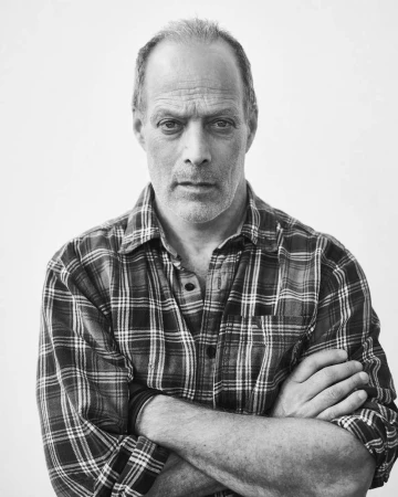 Black and white portrait of a middle-aged man in a plaid shirt with crossed arms against a white background in an indoor setting.