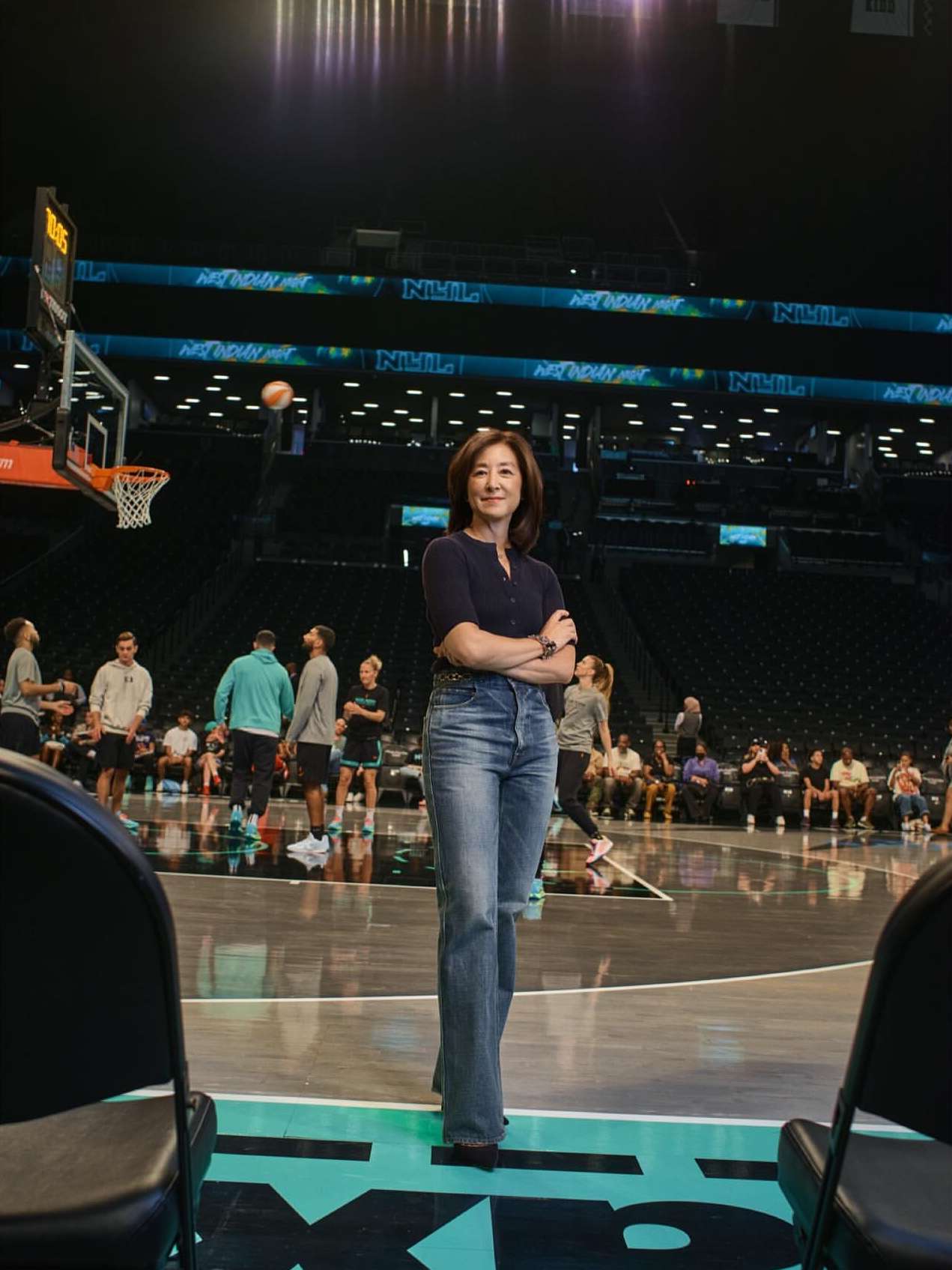 Woman stands confidently on a basketball court in jeans and a navy top, with players and spectators blurred in the background.