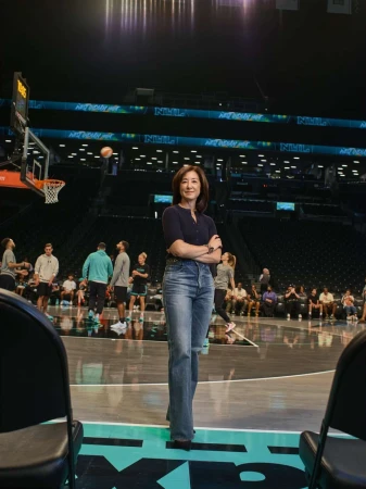 Woman stands confidently on a basketball court in jeans and a navy top, with players and spectators blurred in the background.