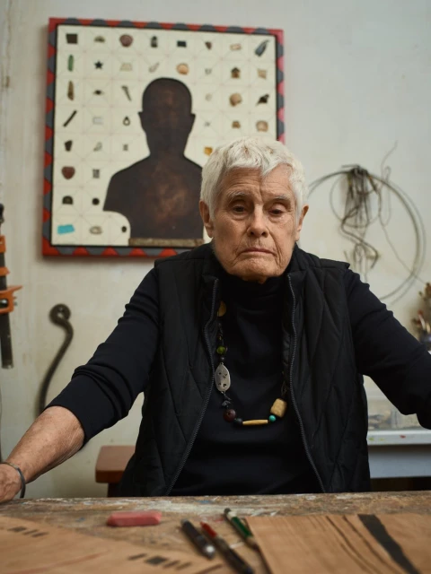 Portrait of an elderly woman with short white hair and a black vest, seated at a table in front of her artwork and an art studio setting.