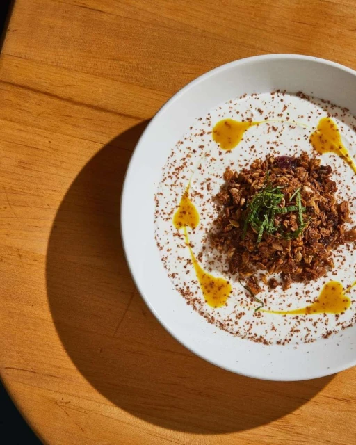 Overhead shot of a white bowl filled with a creamy base, topped with granola, herbs, and golden drizzle on a wood table.