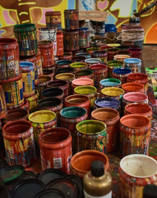 A large collection of open, used paint cans displaying a vibrant array of colors, stacked tightly on a table with paint-splattered surfaces.